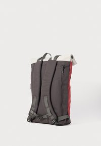 Ryggsekk med rull-topp design, laget av grått og rødt stoff, med justerbare stropper, en sidelomme med glidelås, og en teksturert overflate.