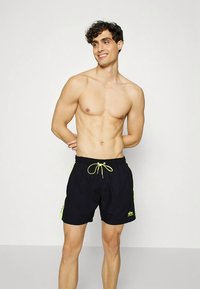 Muž bez trička s kučeravými tmavými vlasmi, oblečený v čiernych plavkách so neonovo žltými pruhmi po stranách a s tkaničkou, usmievajúci sa a hľadiaci do strany.