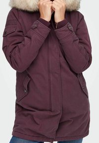 Kurtka parka w kolorze bordo z kapturem wyściełanym futrem, z zamkiem błyskawicznym z przodu, dwoma skośnymi kieszeniami oraz regulowanymi mankietami. Gładka tekstura tkaniny.