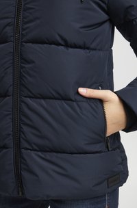 Giacca imbottita blu marina con sezioni trapuntate, chiusura con zip e tasche laterali. Presenta un esterno liscio e resistente all'acqua e polsini elasticizzati.