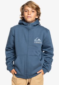 Sweat à capuche zippé bleu avec un logo à l'avant, poches kangourou et une texture douce. Il dispose d'une capuche ajustable et de détails de couture.