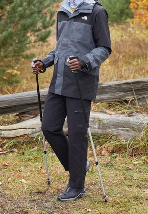 Osoba ubrana w szary i czarny strój outdoorowy, trzymająca dwie kijki trekkingowe, stojąca na trawiastej ścieżce obok drewnianego ogrodzenia i jesiennych liści.