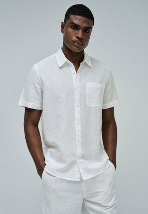 Camisa de lino blanca de manga corta con cuello y un bolsillo en el pecho, que presenta un cierre frontal de botones y un corte relajado. Textura suave en general.