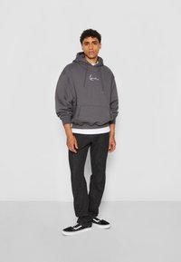 Hettegenser i mørkegrå bomull med lomme på ryggen og hvit skriptlogo. Kombinert med svarte jeans og svart-hvite sneakers.