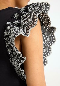 Sort top med intrikate blondeflæser og hvid broderi. Nærbillede af tekstur og detaljerede blomstermønstre langs kanterne.