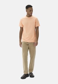 T-shirt en coton orange clair assorti à un pantalon beige et des chaussures noires. Le modèle se tient légèrement tourné à gauche, les mains détendues sur les côtés.