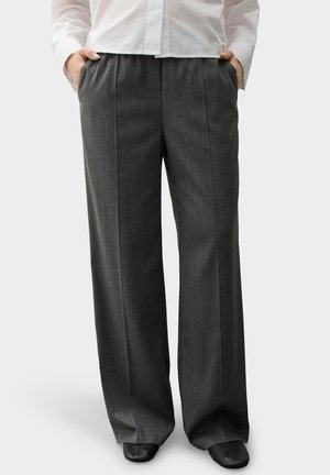 Personne portant un pantalon large gris foncé et des chaussures noires, les mains dans les poches, vêtue d'une chemise blanche sur un fond uni.