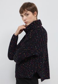 Un maglione nero chunky con colletto alto, caratterizzato da spruzzi multicolori. Il design include una vestibilità ampia e spacco laterale all'orlo.
