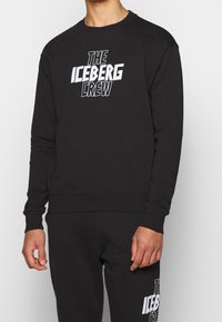 Felpa nera a girocollo in cotone, con un grande testo grafico bianco "THE ICEBERG CREW" sulla parte frontale. Maniche e orlo a coste.