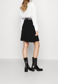 Femme debout, portant une jupe noire mi-longue avec une ceinture "HUGO", une chemise blanche à manches longues et des bottes noires mi-mollet à talons blocs.