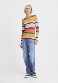 Haut à manches longues à rayures multicolores en tissu côtelé, présentant des teintes de jaune, rouge, bleu et vert, associé à un jean en denim bleu clair.
