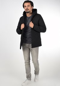 Jachetă neagră cu glugă, cu închidere prin fermoar, având un finisaj neted. Asortată cu blugi gri și adidași albi, purtată de un model masculin în picioare.