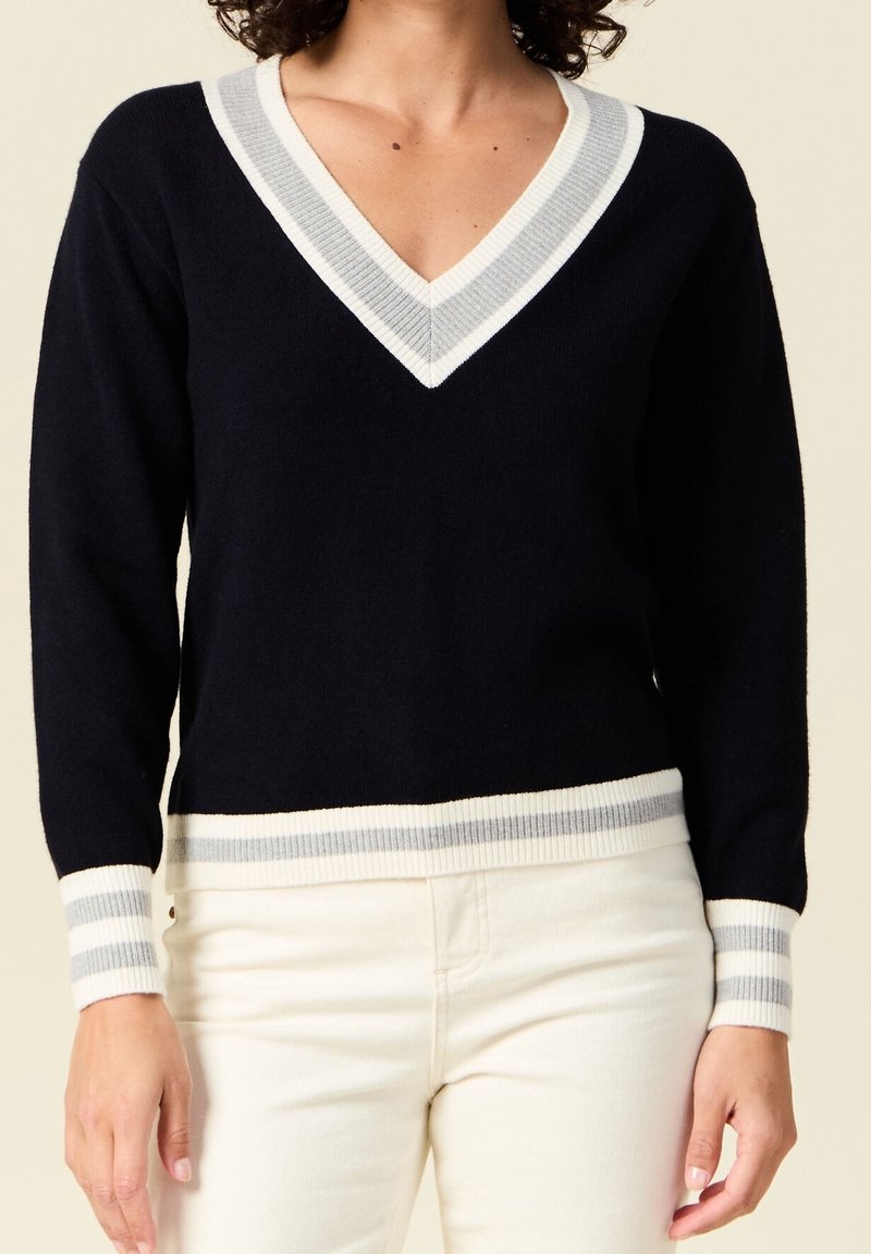 Femme portant un pull noir en V avec un bord rayé blanc et gris sur le col, les poignets et l'ourlet, associé à un pantalon écru.