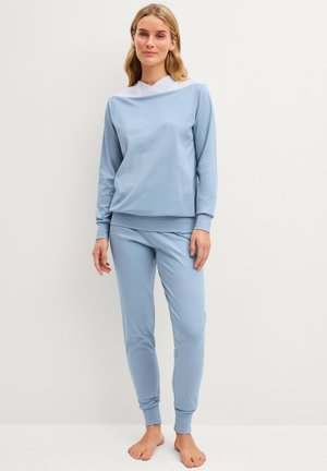 Jonge vrouw staat blootsvoets en draagt een lichtblauw top met lange mouwen en een broek, tegen een effen witte achtergrond.