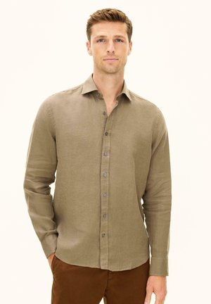 Mies, jolla on lyhyet hiukset, pukeutuneena pitkähihaiseen beigenväriseen nappipaitaan ja ruskeisiin housuihin, seisoo yksiväristä vaaleaa taustaa vasten.