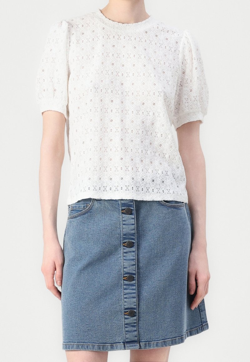 Blouse blanche en dentelle à manches courtes associée à une jupe en denim bleu à boutons devant, portée par une personne debout sur un fond uni.