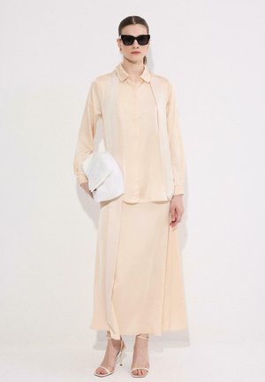 Γυναίκα που φορά εκρού μπλούζα με μακριά μανίκια και φούστα, κρατά λευκή τσάντα χειρός, φορά μαύρα γυαλιά ηλίου και μπεζ σανδάλια με τακούνι, στέκεται μπροστά σε λευκό τοίχο.