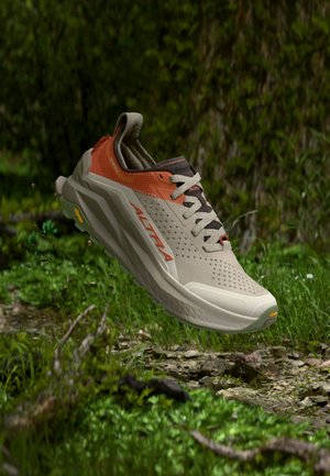 Laufschuh in der Luft über einem grasbewachsenen Waldboden, beige und orange mit sichtbarem "ALTRA"-Logo und Vibram-Sohle.