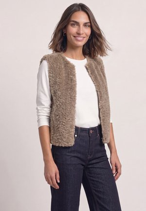 Femme souriante portant une veste beige duveteuse sur une chemise blanche à manches longues et un jean taille haute foncé sur un fond uni.