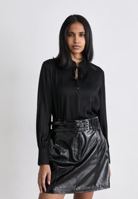 Blusa nera in seta con dettaglio al collo a fiocco, abbinata a una mini gonna in pelle nera con cintura a doppia fibbia e texture visibile.