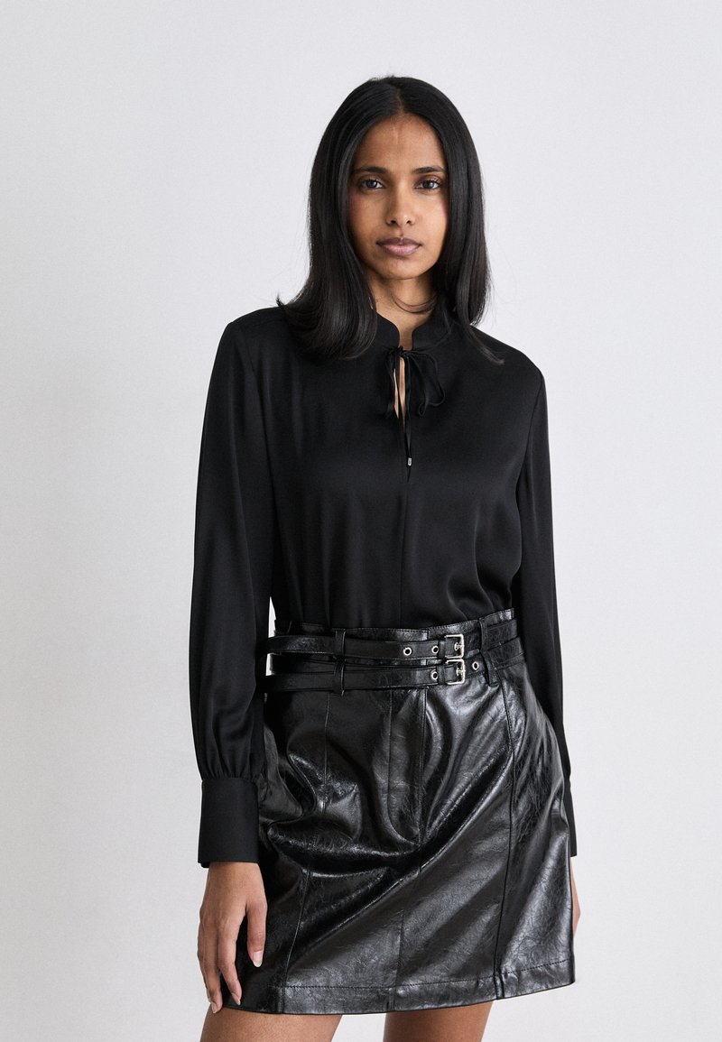 Blusa nera in seta con dettaglio al collo a fiocco, abbinata a una mini gonna in pelle nera con cintura a doppia fibbia e texture visibile.