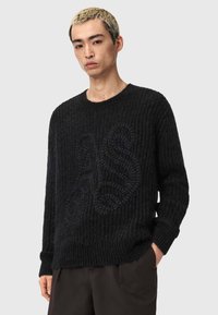 Zwarte gebreide trui met ribbeltextuur, voorzien van een monochrome reliëflogo. Ronde hals en lange mouwen, gecombineerd met donkere broeken.