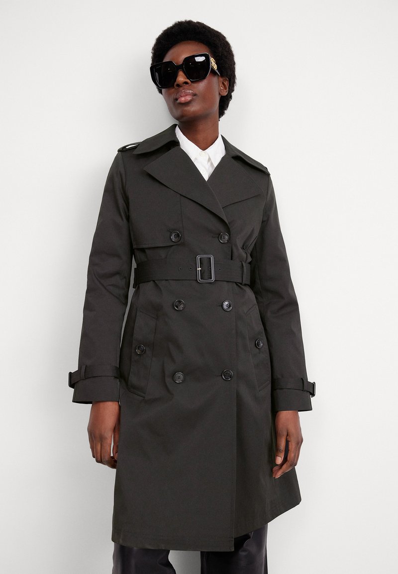 Dunkelgrüner, doppelt behandelter Trenchcoat mit einem Gürtel, Schulterklappen und zwei Vorderaschen. Das Modell trägt übergroße schwarze Sonnenbrillen.