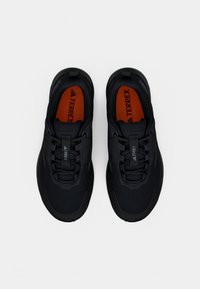 Schwarze Sportschuhe mit einem Mesh-Obermaterial, strukturierten Overlays und einer Gummisohle. Die Innensohlen sind orange mit dem sichtbaren "TERREX"-Logo.