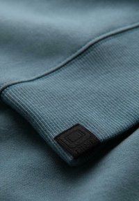 Tela gris-azul con un patrón ribeteado texturizado y una etiqueta de marca negra. El material es suave y liso, mostrando pliegues y contornos.