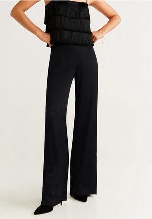 Frau trägt schwarze Hosen mit weitem Bein und ein ärmelloses, geschichtetes Fransen-Top, steht mit den Händen in den Hüften vor einem schlichten Hintergrund.