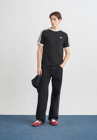 Čierne bavlnené tričko s bielym lemom a tromi bielymi pruhmi na rukávoch, skombinované s voľnými čiernymi džínsy a červenými športovými topánkami.