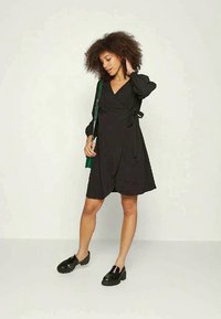 Vestido preto envelopado com mangas longas, apresentando um cinto à cintura. A modelo usa sapatos de plataforma pretos e segura uma bolsa verde. Tecido suave.