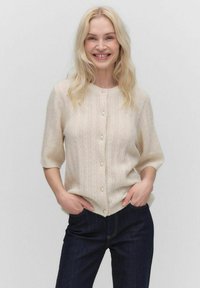 Lichtbeige cardigan met een textuurpatroon, korte mouwen en parelknopen, gedragen over donkere spijkerbroeken. Zacht gebreid materiaal.