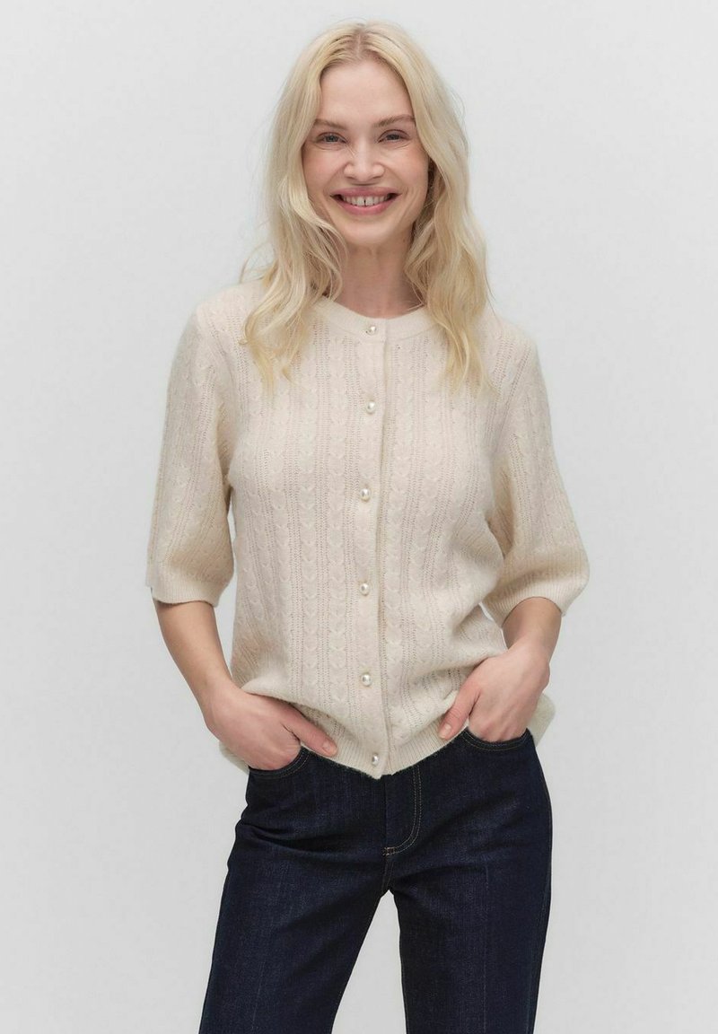 Lichtbeige cardigan met een textuurpatroon, korte mouwen en parelknopen, gedragen over donkere spijkerbroeken. Zacht gebreid materiaal.