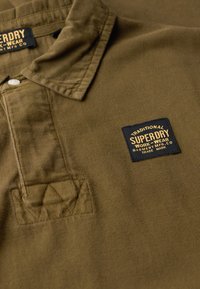 Braun gestreiftes Polohemd mit einem schwarzen Etikett und goldener Schrift. Der Kragen ist strukturiert, und die Brusttasche zeigt ein Rautenmuster.