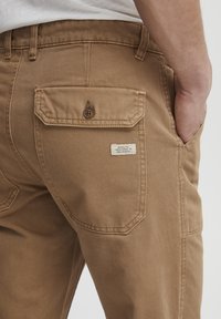 Tan broek van een gestructureerde, duurzame stof. Heeft een achterzak met knoopsluiting en een genaaid label met informatie over het onderhoud van de stof.