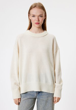 Lichtcrème oversized trui met geribbelde kraag en manchetten, gemaakt van getextureerde brei stof. Draagt met high-waisted blauwe denim jeans.