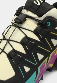 Chaussures de trail légères avec une tige crème, des accents noirs, une semelle texturée pour la traction, un système de laçage élastique et des détails violets et turquoise éclatants.