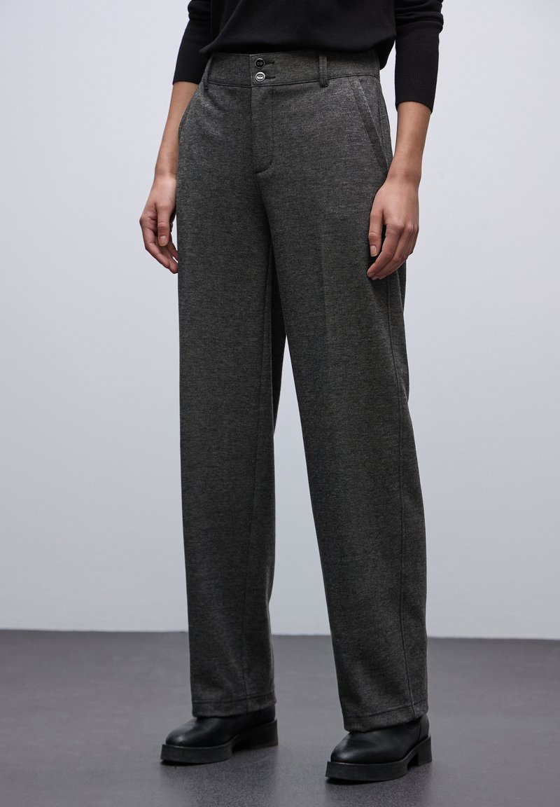 Grijze wijde broek van zacht, getextureerd materiaal. Ze hebben twee knopen en een vlakke voorkant, gecombineerd met zwarte enkellaarzen.
