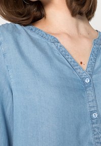 Chemise en denim bleu clair avec un col en V, une patte de boutonnage sur le devant et une texture douce. Détails de couture visibles le long du col et de la patte de boutonnage.