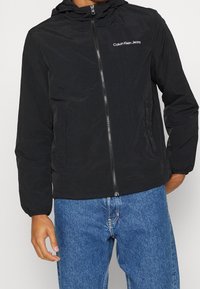 Persona vestida con una chaqueta con capucha de Calvin Klein Jeans negra con cremallera y unos vaqueros azules, de pie frente a un fondo liso.