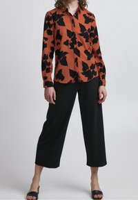 Chemise boutonnée orange brûlé avec motif floral noir, associée à un pantalon noir à jambes larges et des sandales noires à enfiler. Texture du tissu lisse.