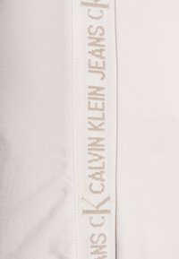 Bílá bavlněná textilie má na jedné straně vertikální pruh s vyšitým nápisem "CALVIN KLEIN JEANS" v světlém béžovém odstínu.