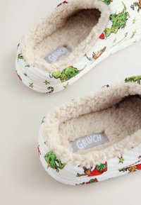 Pantofole accoglienti con un tessuto a strisce bianche, dotate di un morbido rivestimento in pile beige. Decorate con vivaci motivi natalizi a tema Grinch.