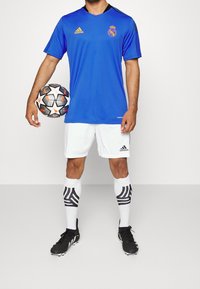 Modrý dres s žlutým logem, bílé šortky, černé kopačky a pruhované ponožky. Drží černo-oranžový fotbalový míč.