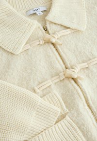 Cream-colored cardigan with a textured knit pattern, featuring a zip front and toggle fasteners. Ribbed cuffs accentuate the design.