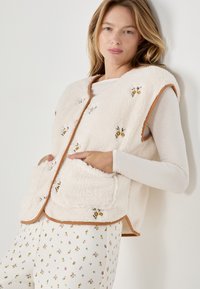Gilet in pile color crema con ricami floreali, rifiniture marroni e tasche laterali, indossato sopra una maglietta bianca a maniche lunghe e pantaloni con fantasia.