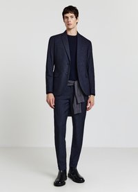 Marineblauer strukturierter Anzug mit einem maßgeschneiderten Blazer und schmal geschnittenen Hosen. Kombiniert mit einem marineblauen Hemd und einem grauen Pullover, der in der Taille gebunden ist. Schwarze Stiefeletten.