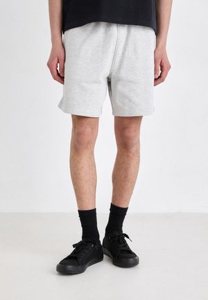 Person trägt hellgraue Shorts, schwarze Socken und schwarze Schnürschuhe und steht auf einem schlichten hellen Boden vor einer weißen Wand.
