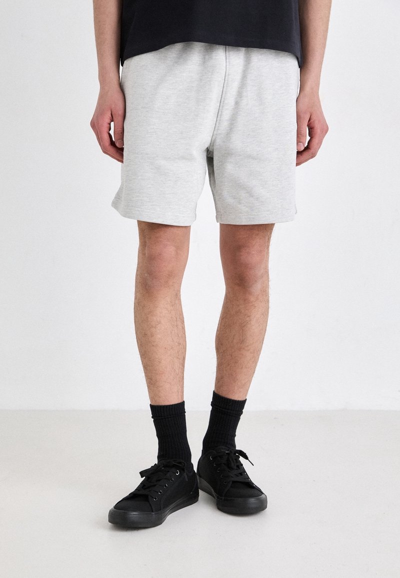 Personne portant un short gris clair, des chaussettes noires et des baskets à lacets noires, debout sur un sol clair uni contre un mur blanc.